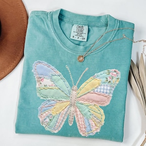May include: A turquoise Comfort Colors t-shirt featuring a patchwork butterfly design in pastel colors. The butterfly wings are made of various patterned fabrics. A gold necklace with a pendant rests on the shirt.