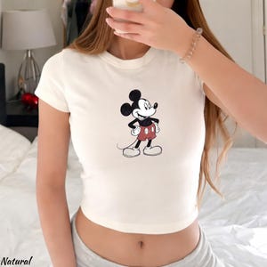 Puede incluir: Top corto color crema con un gr&aacute;fico de Mickey Mouse. El personaje de dibujos animados est&aacute; representado en negro, rojo y blanco. La palabra "Natural" est&aacute; impresa en la esquina inferior izquierda de la imagen.