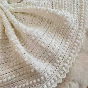 May include: A white, handmade crochet blanket with a textured pattern. The blanket features a combination of raised bobbles and textured stitches, creating a soft and inviting appearance. The edges are finished with a scalloped border.