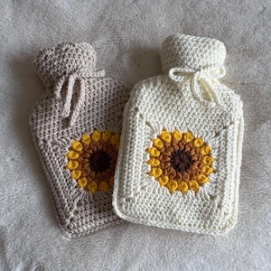 Könnte beinhalten: Zwei gehäkelte Wärmflaschenbezüge. Einer ist beige, der andere cremefarben. Jeder hat ein Sonnenblumenmuster in Gelb und Braun. Die Bezüge haben einen Kordelzugverschluss oben.