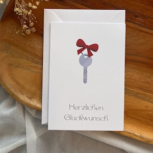 Peut inclure: Une carte de vœux blanche illustrée d'une clé gris clair ornée d'un nœud rouge. Le texte "Herzlichen Glückwunsch" est imprimé en noir sous la clé. La carte est posée sur un plateau en bois avec une branche de fleurs séchées gypsophile.