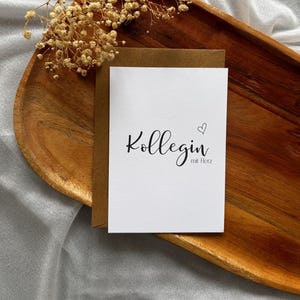 May include: A white greeting card with the word "Kollegin" and "mit Herz" in black script, accompanied by a small heart. The card rests on a brown envelope, placed on a wooden tray with dried flowers.
