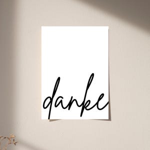 May include: A rectangular white card featuring the word danke written in a black, flowing, cursive script font. The text is positioned in the lower half of the card, with the letters connected in a continuous, handwritten style against a plain background.