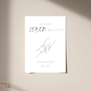Op de afbeelding: Een witte rechthoekige print met een zwarte lijntekening van een volwassen hand die de hand van een klein kind vasthoudt. De tekst luidt DU HAST LEBEN BEGLEITET en DANKE FÜR ALLES in een strak lettertype, met LEBEN in een cursieve stijl.