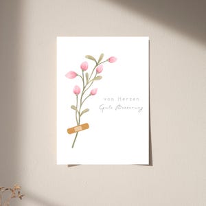 Könnte beinhalten: Eine weiße Grußkarte mit einer Aquarellillustration eines grünen Stiels mit rosa Blütenknospen und einem beigen Pflaster, das um den unteren Stiel gewickelt ist. Der Text auf der Karte lautet von Herzen Gute Besserung in schwarzer Schrift.
