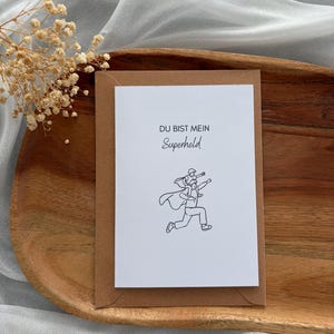 May include: A white greeting card with a brown envelope. The card features a black line drawing of a person carrying another person, with the words "DU BIST MEIN Superheld" in black text.