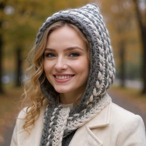 Könnte beinhalten: Eine Frau trägt ein gehäkeltes Kapuzen- und Schal-Set in Grau und Weiß. Die Kapuze ist hochgezogen und rahmt das Gesicht ein, und der Schal ist um den Hals drapiert. Die Frau trägt auch einen cremefarbenen Mantel.