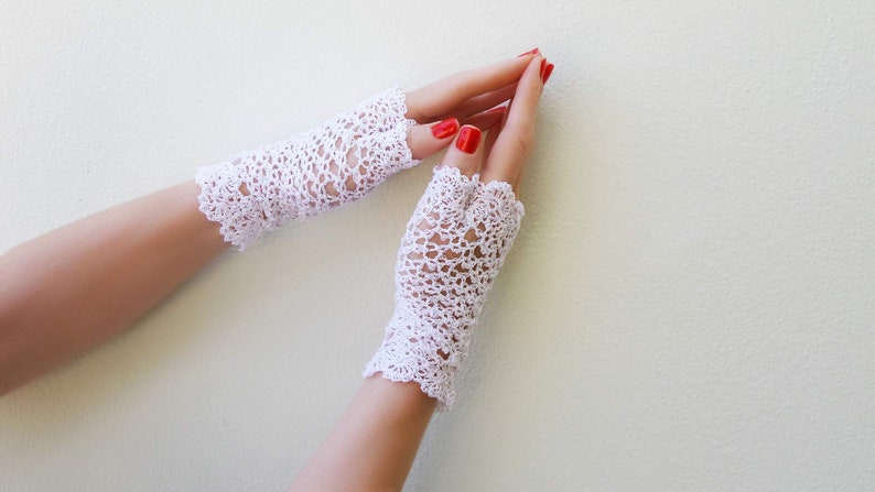May include: A pair of white crocheted fingerless gloves with a delicate lace pattern. The gloves are worn on a person's hands with red nail polish.