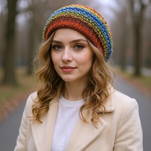 May include: A colorful, knitted beanie hat with a rainbow pattern. The hat features shades of blue, green, yellow, orange, and red. The hat is worn by a person with long, wavy blonde hair, wearing a white shirt and a cream-colored coat.