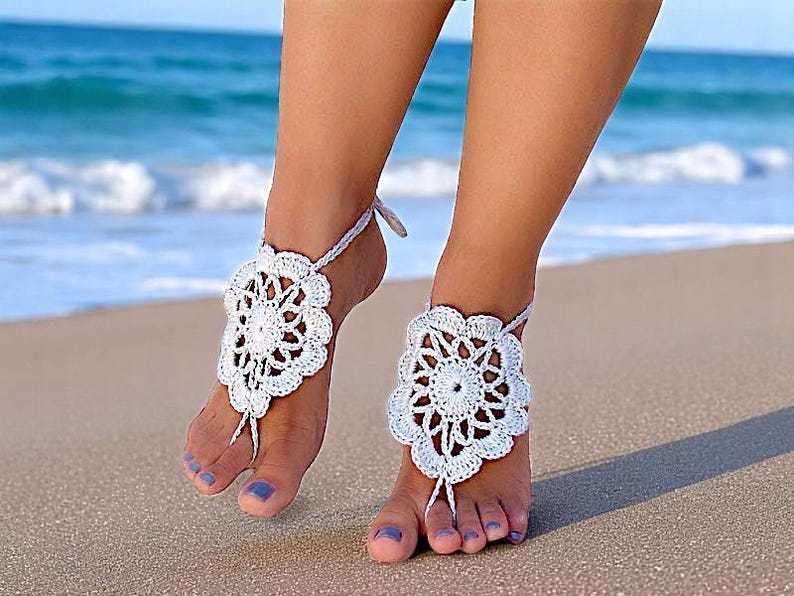 May include: White crochet barefoot sandals with a floral design. The sandals are worn on a woman's feet on a sandy beach.