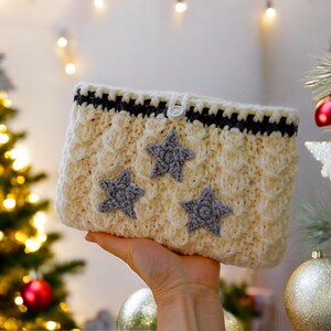 Puede incluir: Una bolsa de ganchillo blanca con ribete negro y tres estrellas grises como adorno. La bolsa tiene un cierre de bot&oacute;n.