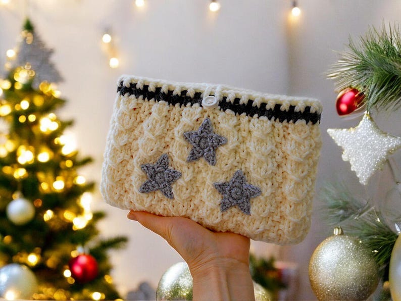 Puede incluir: Una bolsa de ganchillo blanca con ribete negro y tres estrellas grises como adorno. La bolsa tiene un cierre de bot&oacute;n.