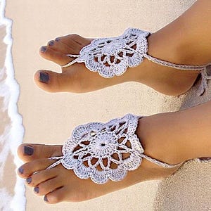 May include: A pair of white crocheted barefoot sandals with a delicate floral design. The sandals are worn on a person's feet, with the toes and ankles visible.