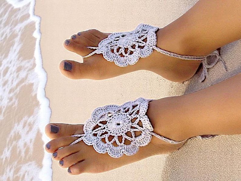 May include: A pair of white crocheted barefoot sandals with a delicate floral design. The sandals are worn on a person's feet, with the toes and ankles visible.
