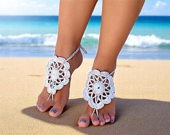 May include: White crochet barefoot sandals with a floral design. The sandals are worn on a woman's feet on a sandy beach.