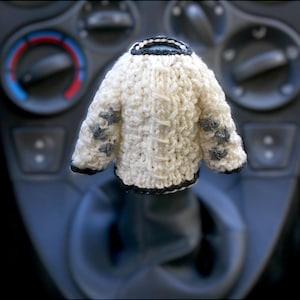 Puede incluir: Un suéter de punto blanco con ribetes negros y un patrón de copo de nieve gris. El suéter está colgado de la palanca de cambios de un coche.