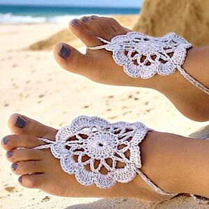 May include: A pair of white crocheted barefoot sandals with delicate floral designs. The sandals are worn on a person's feet on a sandy beach.