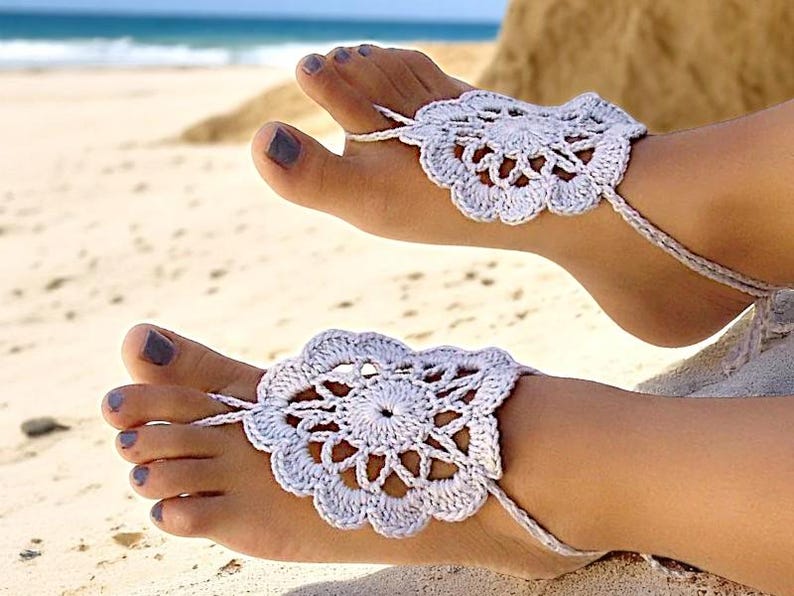 May include: A pair of white crocheted barefoot sandals with delicate floral designs. The sandals are worn on a person's feet on a sandy beach.