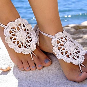 May include: White crocheted barefoot sandals with a flower design. The sandals are worn on a woman's feet.