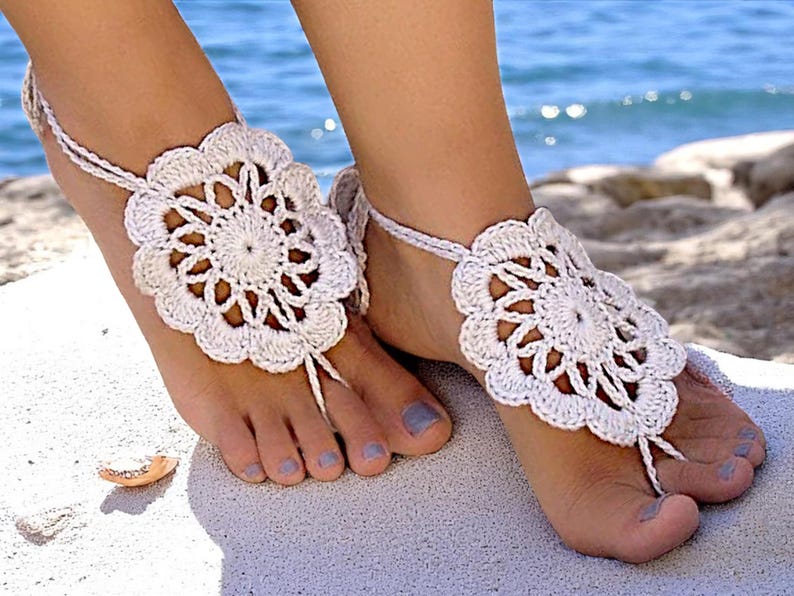 May include: White crocheted barefoot sandals with a flower design. The sandals are worn on a woman's feet.