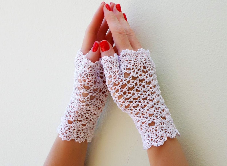 May include: A pair of white crocheted fingerless gloves with a delicate lace pattern. The gloves are worn on a person's hands with red nail polish.