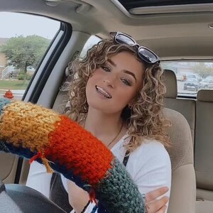 May include: A woman with curly blonde hair is wearing sunglasses and a white shirt. She is sitting in the driver's seat of a car and has a colourful knitted steering wheel cover.