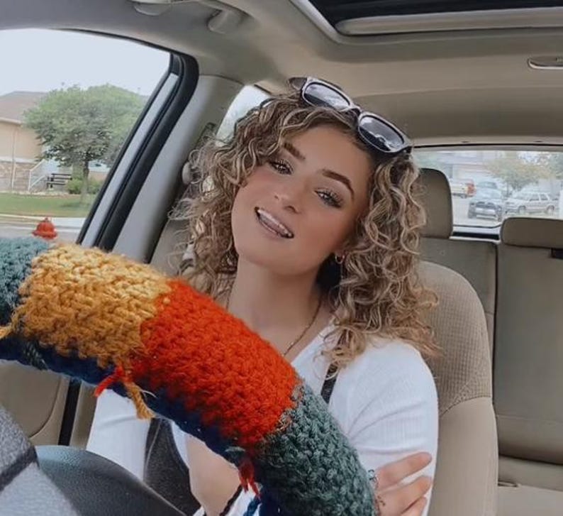 May include: A woman with curly blonde hair is wearing sunglasses and a white shirt. She is sitting in the driver's seat of a car and has a colourful knitted steering wheel cover.