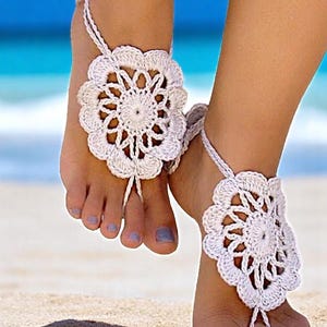 May include: White crocheted barefoot sandals with a flower design. The sandals are worn on a woman's feet on a sandy beach.