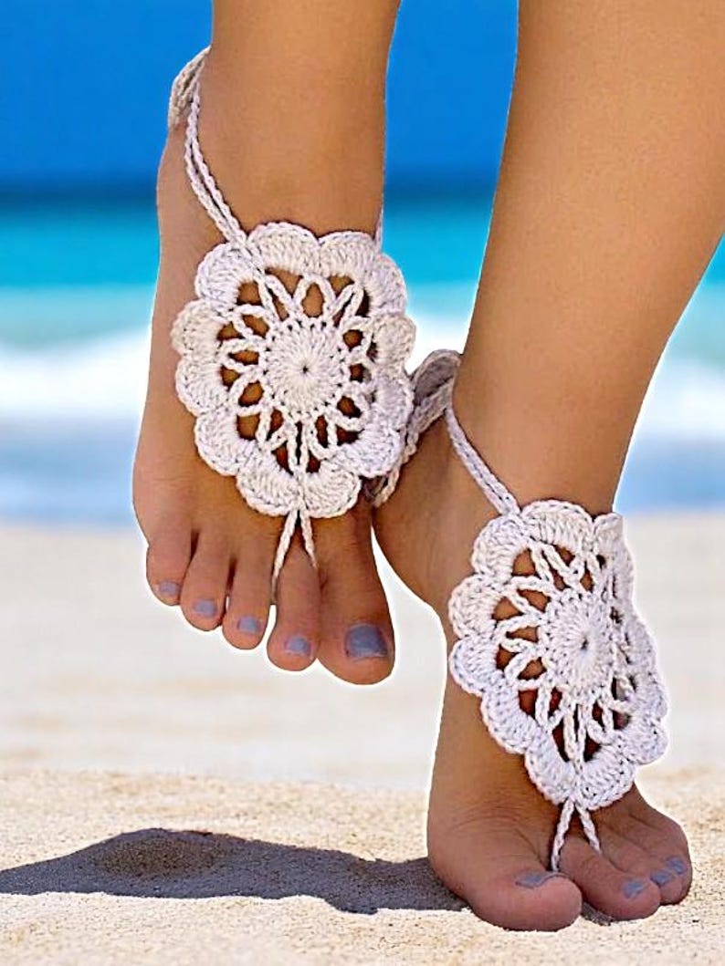 May include: White crocheted barefoot sandals with a flower design. The sandals are worn on a woman's feet on a sandy beach.