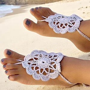 May include: A pair of white crocheted barefoot sandals with a delicate floral design. The sandals are tied around the ankles and have a tassel detail.