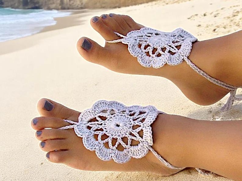 May include: A pair of white crocheted barefoot sandals with a delicate floral design. The sandals are tied around the ankles and have a tassel detail.