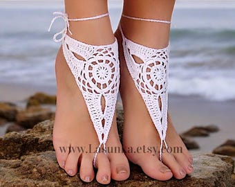 May include: White crochet barefoot sandals with a floral design. The sandals are tied around the ankle with a thin white cord. The image shows the sandals on a person's feet, with a beach background.