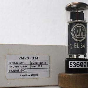 Könnte beinhalten: Eine Valvo EL34 Vakuumröhre mit klarem Glaskolben und schwarzen internen Komponenten, versehen mit dem Valvo-Logo und der Aufschrift EL 34. Sie steht auf einem Holzsockel neben einer weißen Schachtel mit einem Etikett, das technische Daten anzeigt.