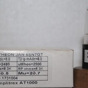 Pode incluir: Um tubo de vácuo Raytheon JAN 6SN7GT com uma base preta rotulada 1731004, posicionado ao lado de uma caixa branca. O rótulo da caixa exibe especificações técnicas, incluindo valores de T1 e T2 Ip mAdc, uMhos, RP ohms, Mu e o número 1731004.