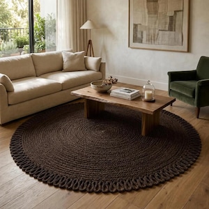 May include: A large, circular, dark brown rug featuring a thick, braided texture and a decorative looped edge. The rug is crafted from a coarse, natural-looking fiber and is positioned on a light-colored wooden floor in a living room setting.