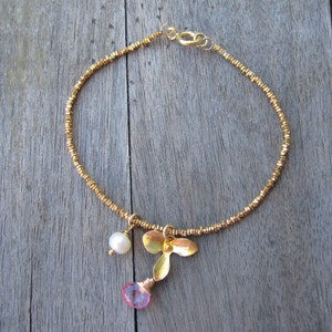 May include: A gold bracelet with a delicate chain and a charm featuring a pink gemstone, a gold flower, and a white pearl.