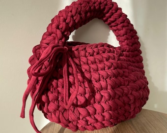 May include: A handmade, red crochet bag with a rounded shape and a sturdy handle. The bag features a textured stitch pattern and a drawstring closure. The bag is made of a soft, thick yarn and is suitable for carrying everyday items.