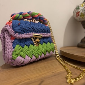 May include: A colorful, handmade crochet handbag with a gold chain strap. The bag features a braided handle and a gold clasp. The body of the bag is a mix of blue, green, pink, and purple crochet.