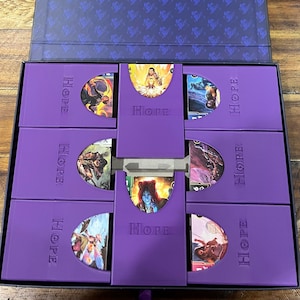 May include: A purple box with a dark blue interior, open on a wooden surface. Inside, there are several purple card holders, each labelled "HOPE." The holders contain illustrated cards. The box is designed for card storage.