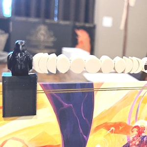 May include: A game board with a colourful illustration, featuring a raven figurine on each end. A series of white, circular game pieces are strung along a metal rod above the board. The board has a purple and yellow design.