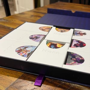 May include: A dark blue box with white card holders, each with a semi-circular cutout displaying a colourful image. The word "Hope" is embossed on each holder. A purple ribbon is attached to the box.