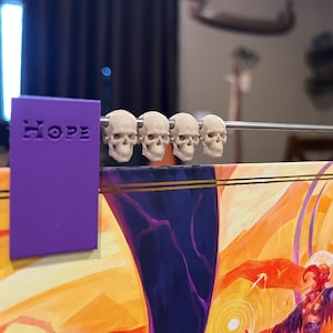 May include: An unusual abacus with a purple frame bearing the words "HOPE" and "Fess". It has small skull beads on a metal rod, set against a colourful, fantasy-themed background.
