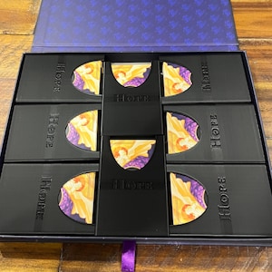 May include: An open, rectangular box with a purple interior and a dark wood exterior. Inside, there are several black boxes with the word "HOPE" printed on them. Each box contains a colourful image with the word "Hope" in the centre.