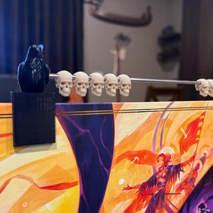 May include: A decorative game board accessory featuring a series of small skull figurines strung on a metal rod. The skulls are cream-colored and are held in place by black raven figurines. The board has a colorful illustration.