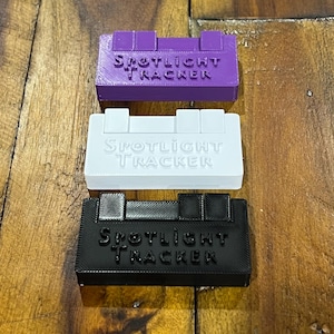 May include: Three rectangular Spotlight Tracker devices in purple, white, and black. Each device has the text "Spotlight Tracker" embossed on the surface. The devices are stacked on a wooden surface.
