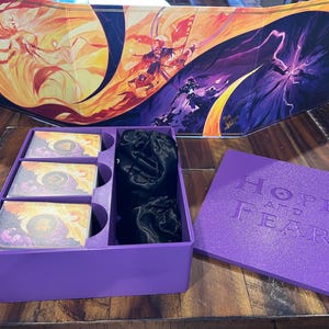 May include: A purple game box with the words "HOPE and FEAR" embossed on the lid. The box contains cards, black drawstring bags, and a game board with a colourful fantasy illustration. A game accessory.