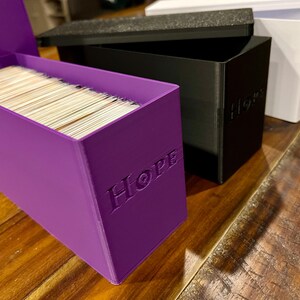 May include: A purple box filled with cards, embossed with the word "HOPE". A black box with "HOPE" and a white box with "Fear" are also visible. The boxes are on a wooden surface.