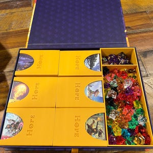 May include: A game box with a purple interior and a yellow tray. The tray holds six yellow boxes labelled "HOPE", and a compartment filled with colourful gems and dice. The box is on a wooden surface.