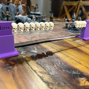 May include: A tabletop game accessory featuring a row of small skull figurines suspended on a metal rod. The purple supports are labeled "Hope" and "Fear". The background includes miniature fantasy figures and scenery.
