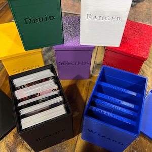 May include: A selection of brightly coloured, 3D-printed card storage boxes. The boxes are labelled with fantasy class names such as "Druid", "Ranger", and "Wizard". The boxes are in various colours, including green, yellow, purple, red, black, and blue. Some boxes contain cards.